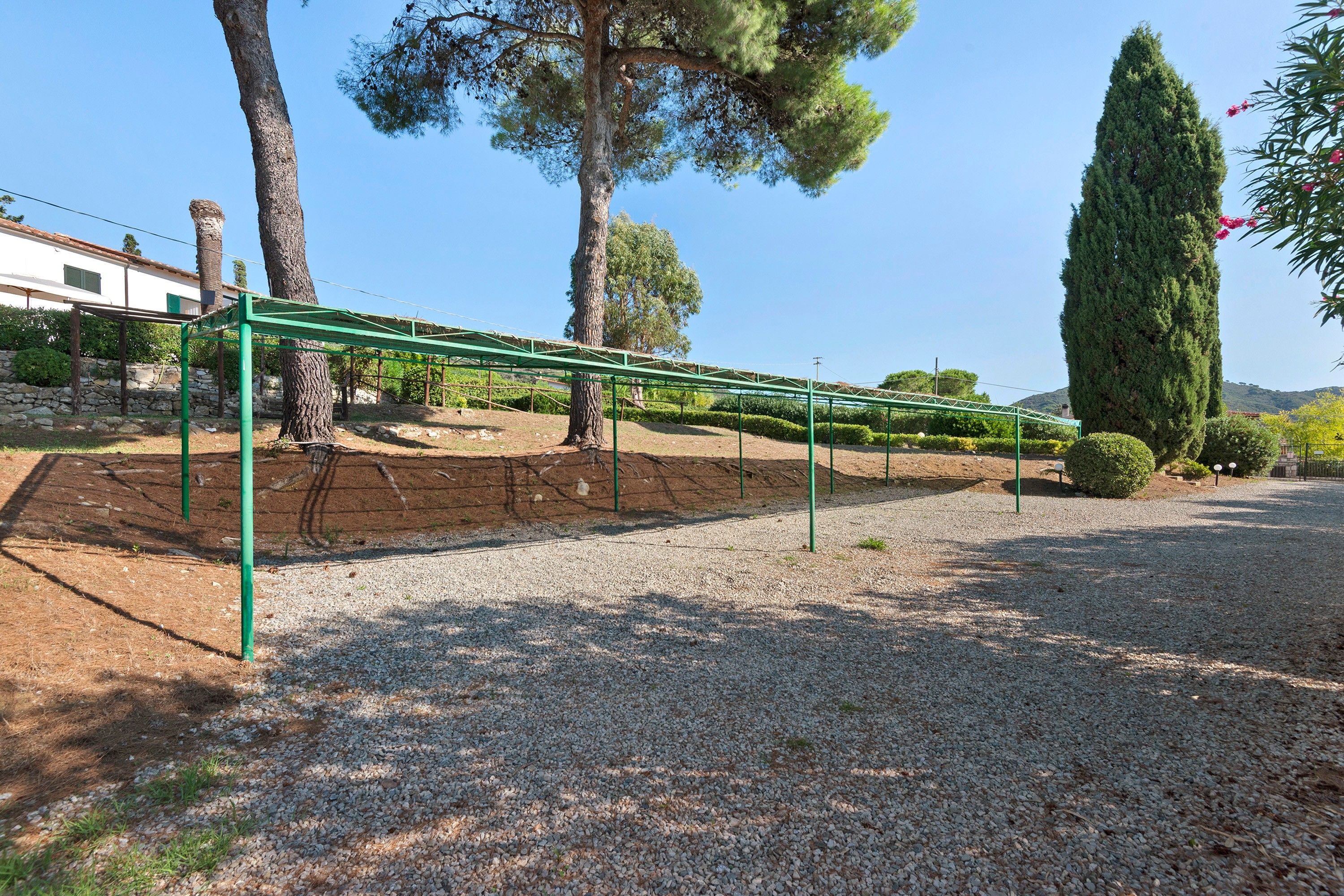 Ein Kiesweg mit einer grünen Metallkonstruktion, vermutlich eine Pergola, umgeben von Bäumen und einer Steinmauer.