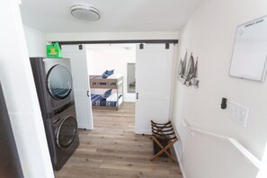 Ein Waschbereich mit Waschmaschine und Trockner, ein Stockbett im Hintergrund und ein Stuhl mit gestreifter Sitzkissen.