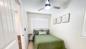 Ein Schlafzimmer mit einem Bett, einem Deckenventilator, einem Fenster mit Jalousien und gerahmten Kunstwerken an der Wand.