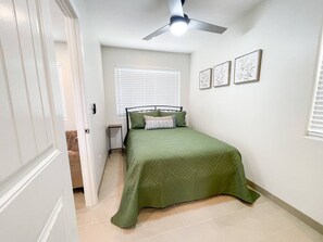Ein Schlafzimmer mit einem Bett, einem Deckenventilator, einem Fenster mit Jalousien und gerahmten Kunstwerken an der Wand.