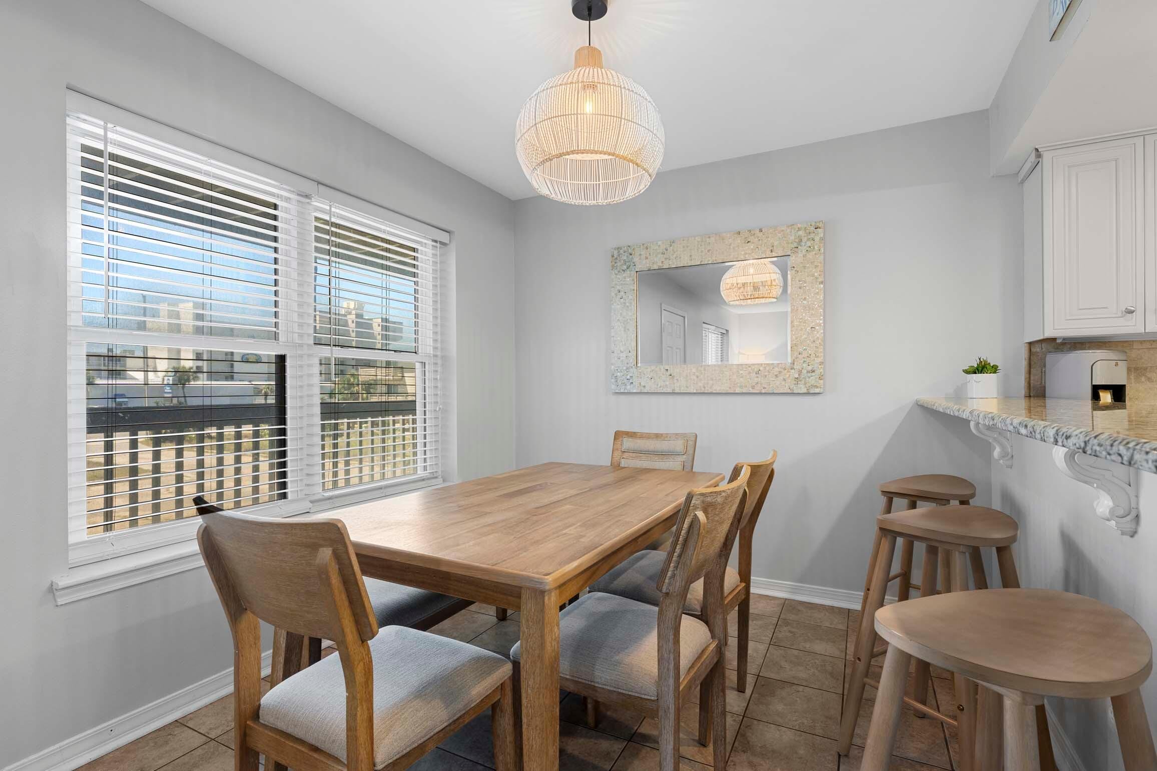 A dining area with a wooden table and chairs, a chandelier, a kitchen counter, and a window with blinds.
