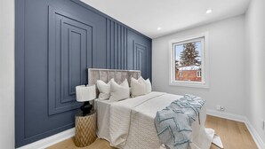 Ein Schlafzimmer mit großem Fenster, einem Bett mit weißer Bettwäsche, einem Nachttisch mit Lampe und einer Wand mit blauem Wandpaneel-Design.
