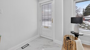 Ein Flur mit einer weißen Tür, einem Fenster mit Blick auf eine verschneite Landschaft, einer Stehlampe und einem Korbwägelchen auf dem Boden.