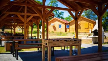 A wooden pavilion with benches and a foosball table, surrounded by a park with a barbecue area and a house in the background.