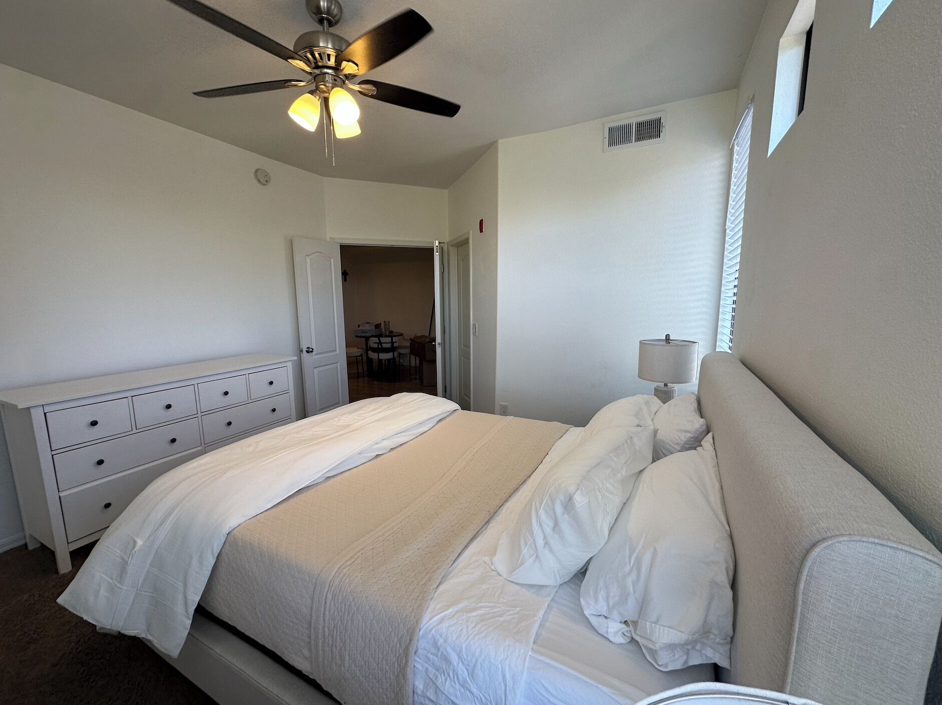 A bedroom with a bed, a ceiling fan, a bedside lamp, and a door leading to a small dining area.