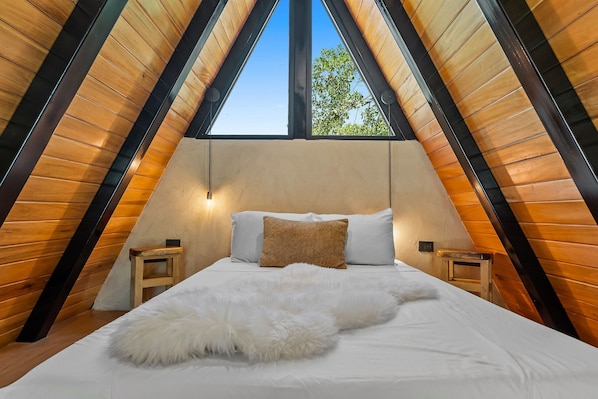 A bedroom with a sloped ceiling, a bed with white bedding, a fluffy white throw, two bedside tables, and a window with a view of trees.
