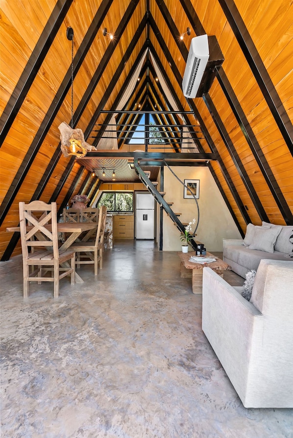 A loft-style room with a sloped ceiling, a dining table with chairs, a sofa, a small kitchen area, and a staircase.