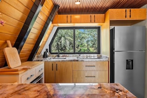A kitchen with wooden cabinets, a stainless steel refrigerator, a marble countertop, and a window with a view of trees.