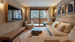 Una sala de estar moderna con un televisor de pantalla plana grande, sofás cómodos, una mesa de centro redonda y un balcón con vistas a árboles cubiertos de nieve.