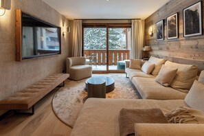 Ein modernes Wohnzimmer mit einem großen Flachbildfernseher, bequemen Sofas, einem runden Couchtisch und einem Balkon mit Blick auf verschneite Bäume.