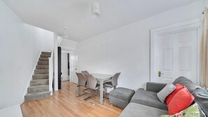 A modern living room with a grey sofa, a dining table with chairs, a staircase, and a white door. - Hounslow Hideaway Cottage (Hounslow)