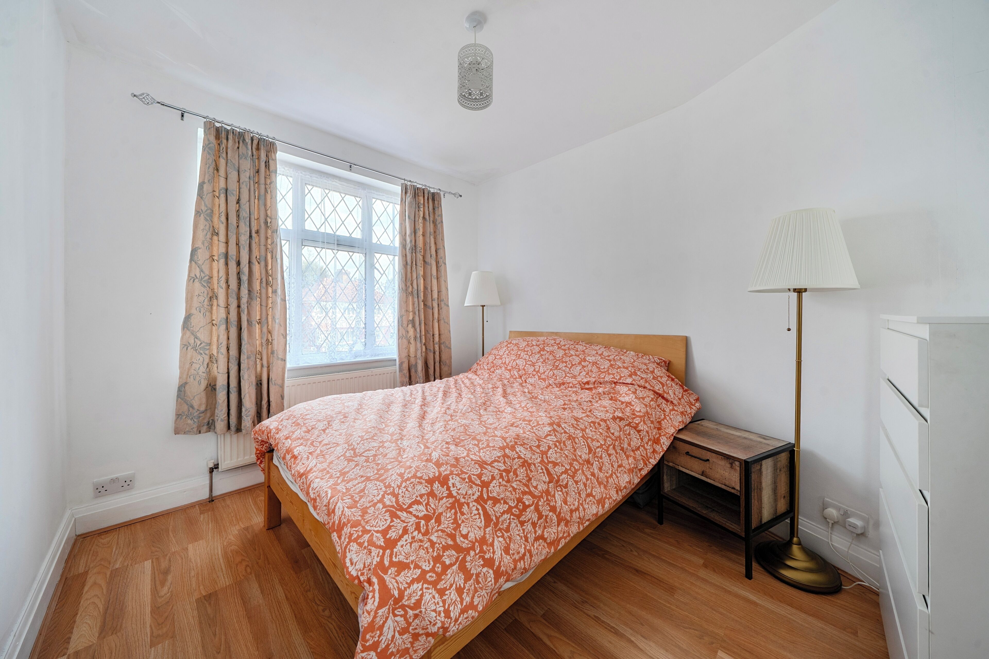 A bedroom with a bed, a bedside table, a lamp, a window with curtains, and a radiator.