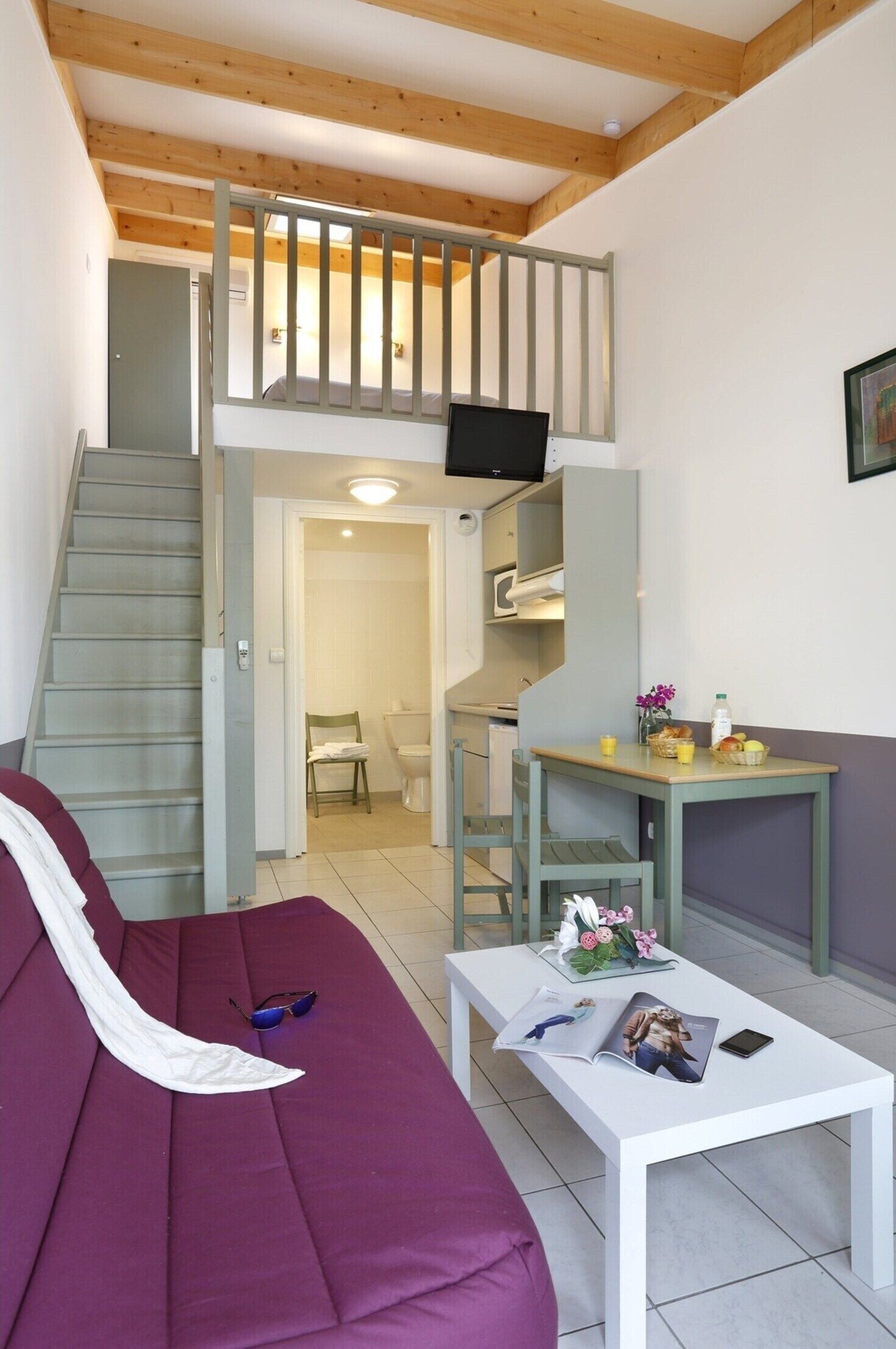 Una sala de estar con un sofá, una mesa de centro con revistas, un televisor en la pared y una escalera que sube a un nivel superior.