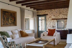 A living room with a sofa, armchair, and a wicker table. There is a painting on the wall and a stone wall with a window. - Son Seriol · Exclusive ECO Villa in Finca w/ views (Inca)