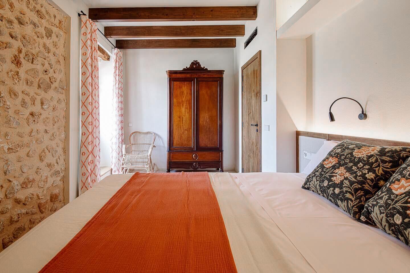 A bedroom with a bed, a wooden wardrobe, a wicker chair, and a patterned pillow.