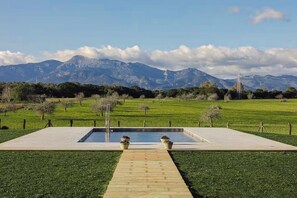 A rectangular pool with two flower pots, surrounded by a paved deck, with a grassy area and trees in the foreground and mountains in the background. - Son Seriol · Exclusive ECO Villa in Finca w/ views (Inca)
