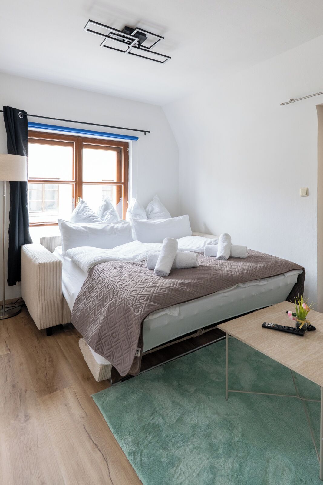 A bedroom with a bed, a bedside table with a remote, a window, and a radiator.