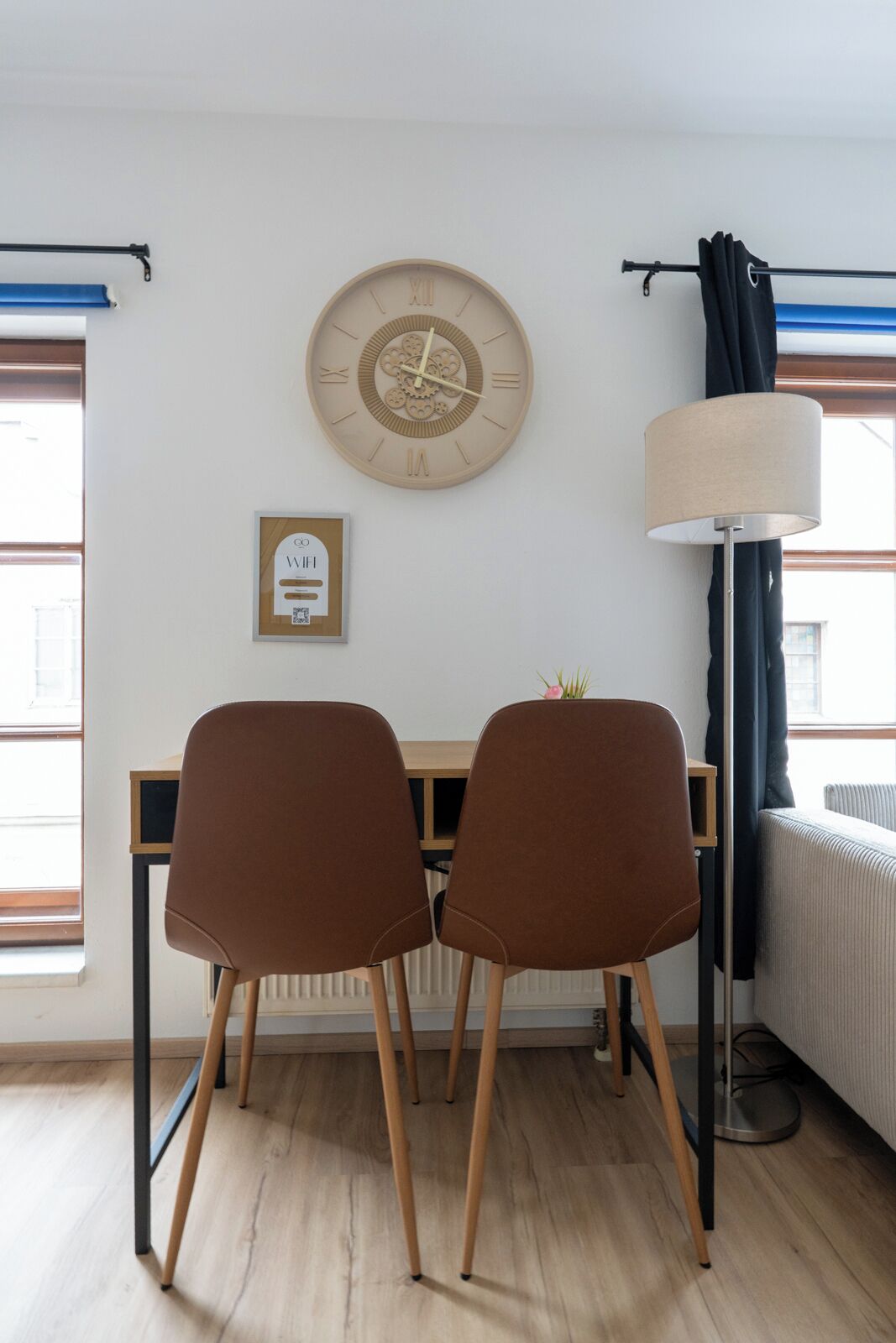 Una stanza con una scrivania in legno, due sedie marroni, una sveglia a muro, una lampada e un segnale WiFi incorniciato.