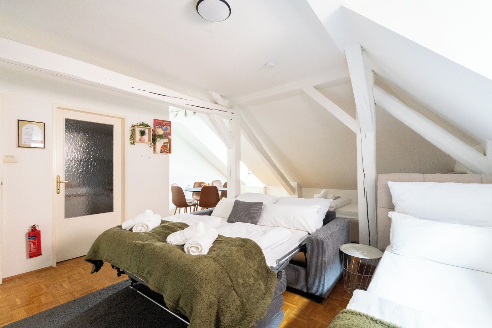 A room with a bed, a small table, and a chair. There is a door with a fire extinguisher on the wall.