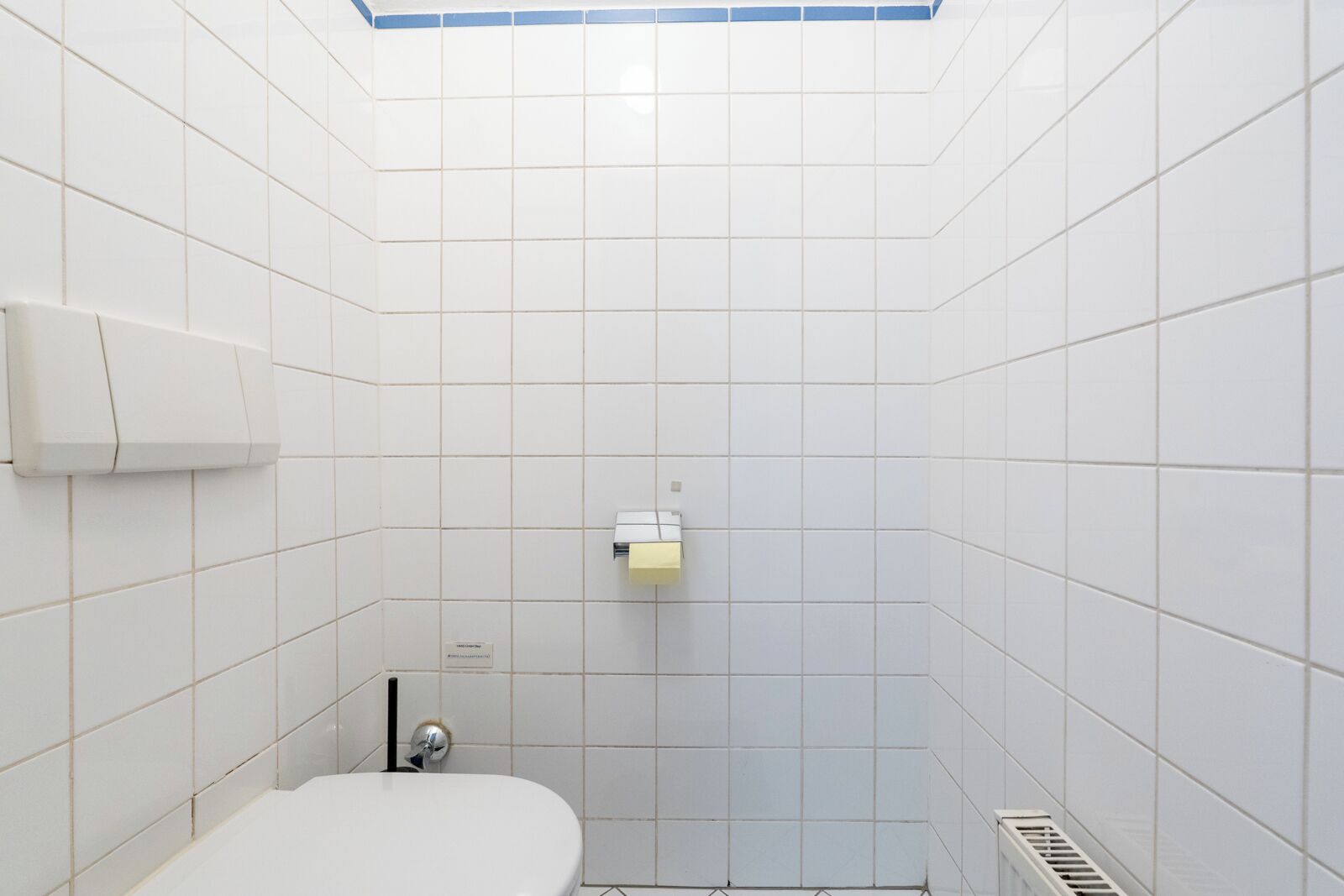 Baño con azulejos blancos, inodoro, dispensador de toallas y rejilla de ventilación.