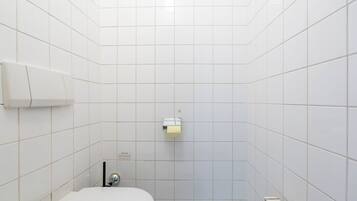 Baño con azulejos blancos, inodoro, dispensador de toallas y rejilla de ventilación.