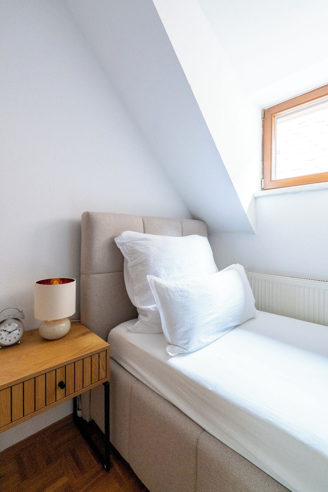 A small, cozy room with a single bed, a nightstand with a lamp, and a window.