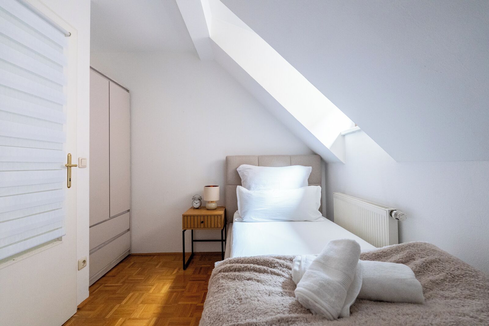 A bedroom with a sloped ceiling, a bed with white bedding, a nightstand with a lamp, and a radiator.