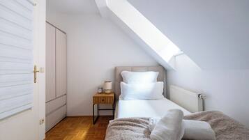 A bedroom with a sloped ceiling, a bed with white bedding, a nightstand with a lamp, and a radiator.