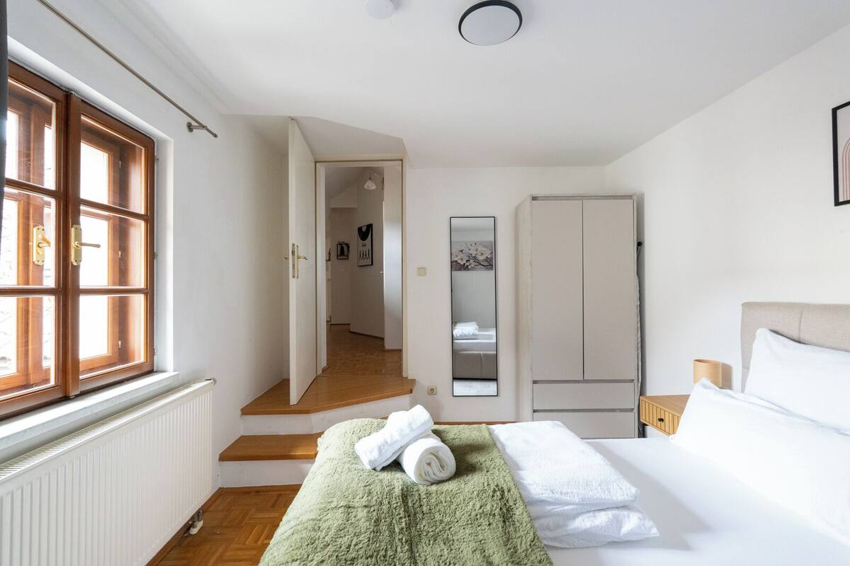 Un dormitorio con una cama, una toalla verde, una toalla blanca, suelo de madera, un radiador, una ventana y una puerta que da a otra habitación.