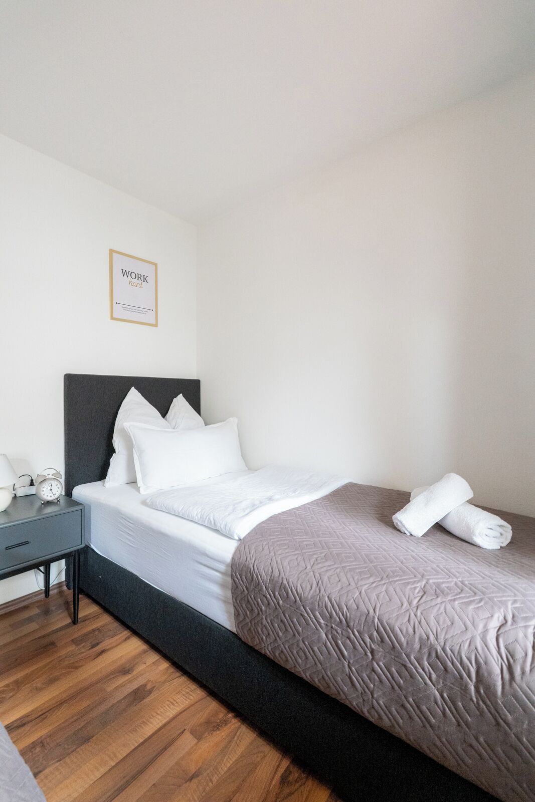 A neatly made bed with white and gray bedding, a bedside table with a lamp and alarm clock, and a framed motivational quote on the wall.