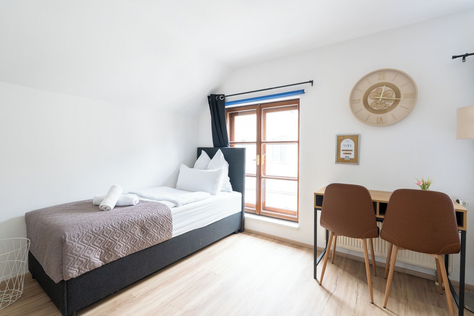 Quarto com cama, escrivaninha, duas cadeiras, janela com cortinas, relógio de parede e radiador.