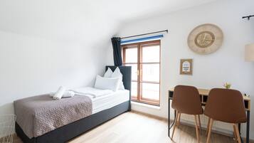 Quarto com cama, escrivaninha, duas cadeiras, janela com cortinas, relógio de parede e radiador.