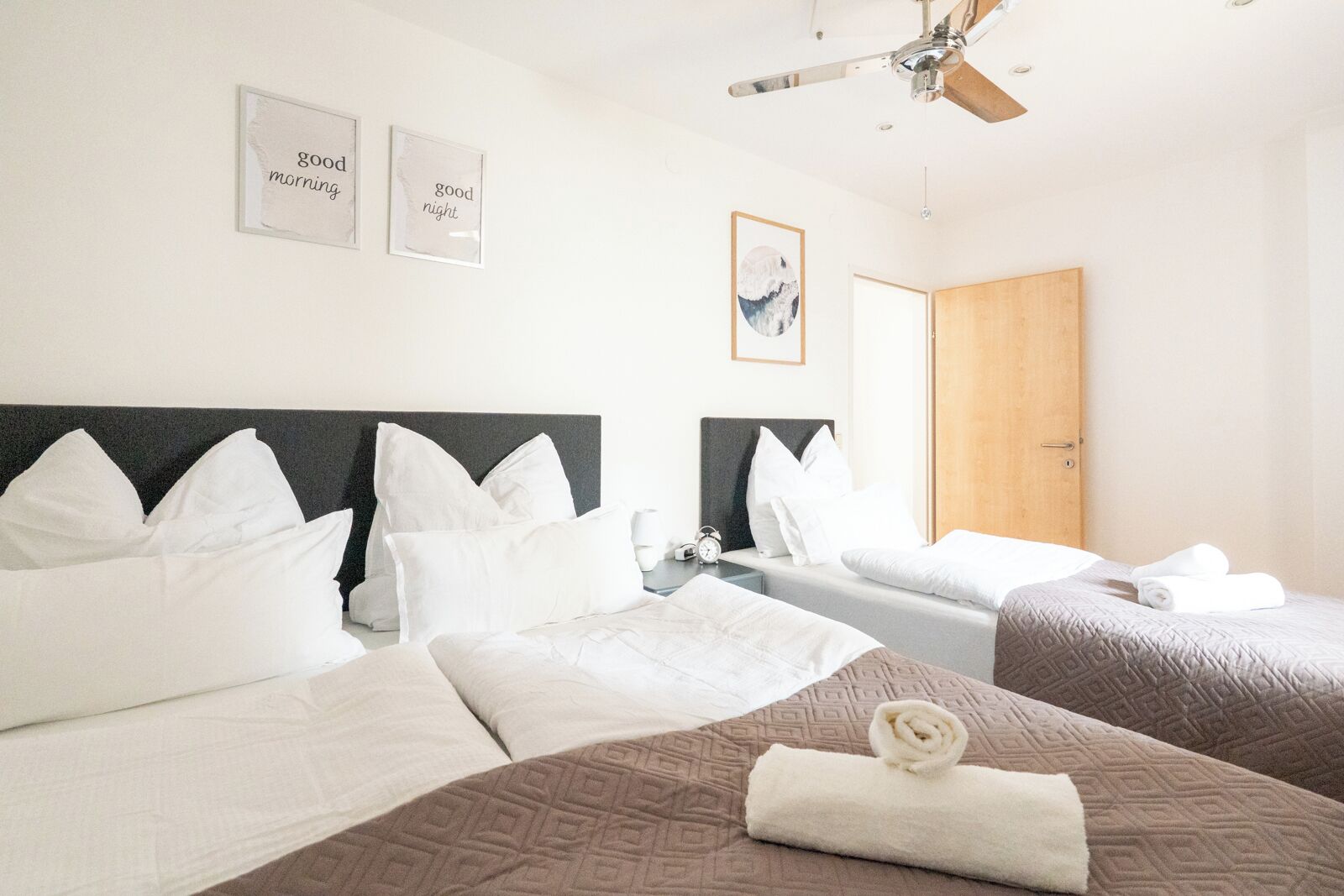 A hotel room with two beds, white bedding, a ceiling fan, a wooden door, and framed pictures on the wall.