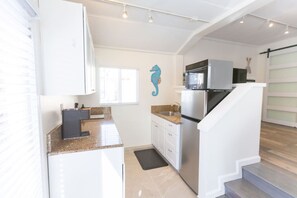 A compact kitchen with a refrigerator, microwave, and stove, a small window, and a staircase leading to another level. - Beachside Bungalow! Steps to Ocean + Bay /Sleeps 6 (San Diego)