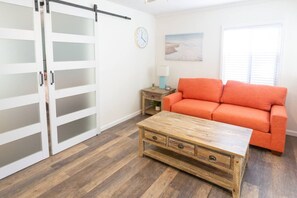 A room with a sliding door closet, a wooden coffee table, an orange sofa, a clock, and a painting on the wall. - Beachside Bungalow! Steps to Ocean + Bay /Sleeps 6 (San Diego)