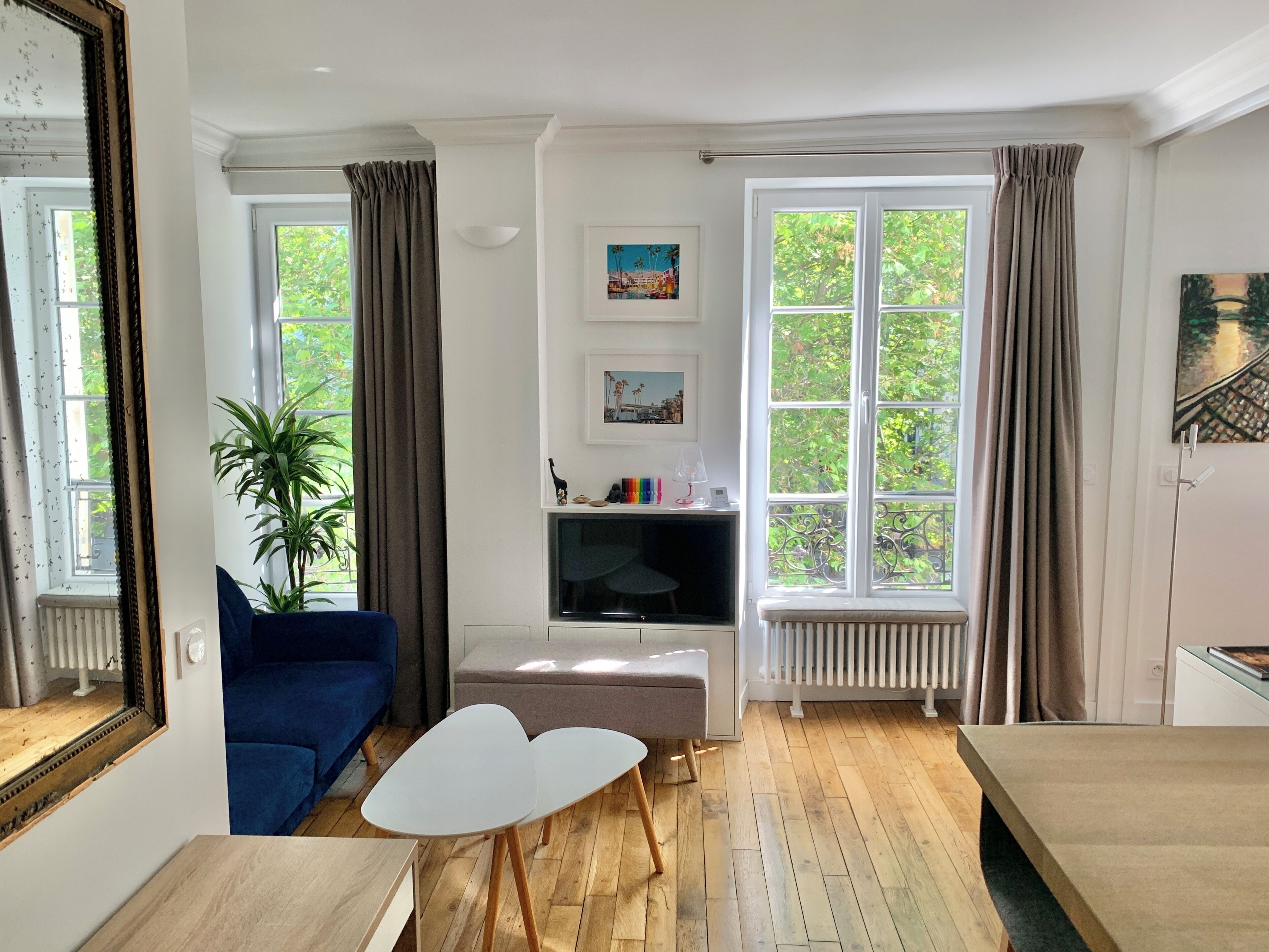 A living room with a blue sofa, a white table, a TV, and a large window with curtains.