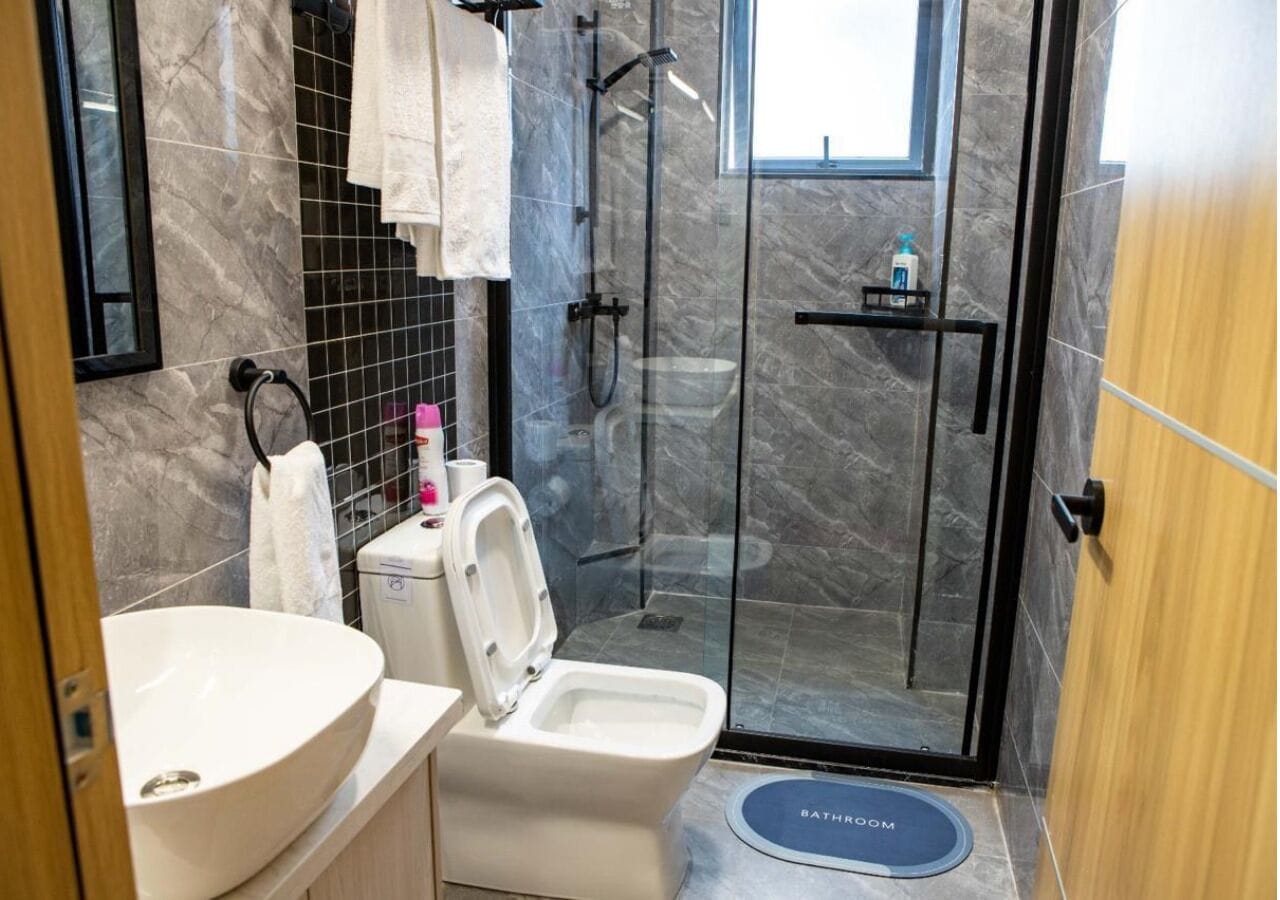 Un baño con lavamanos, inodoro y ducha blancos. Incluye un perchero, un estante pequeño con artículos de aseo y una puerta de ducha de vidrio.