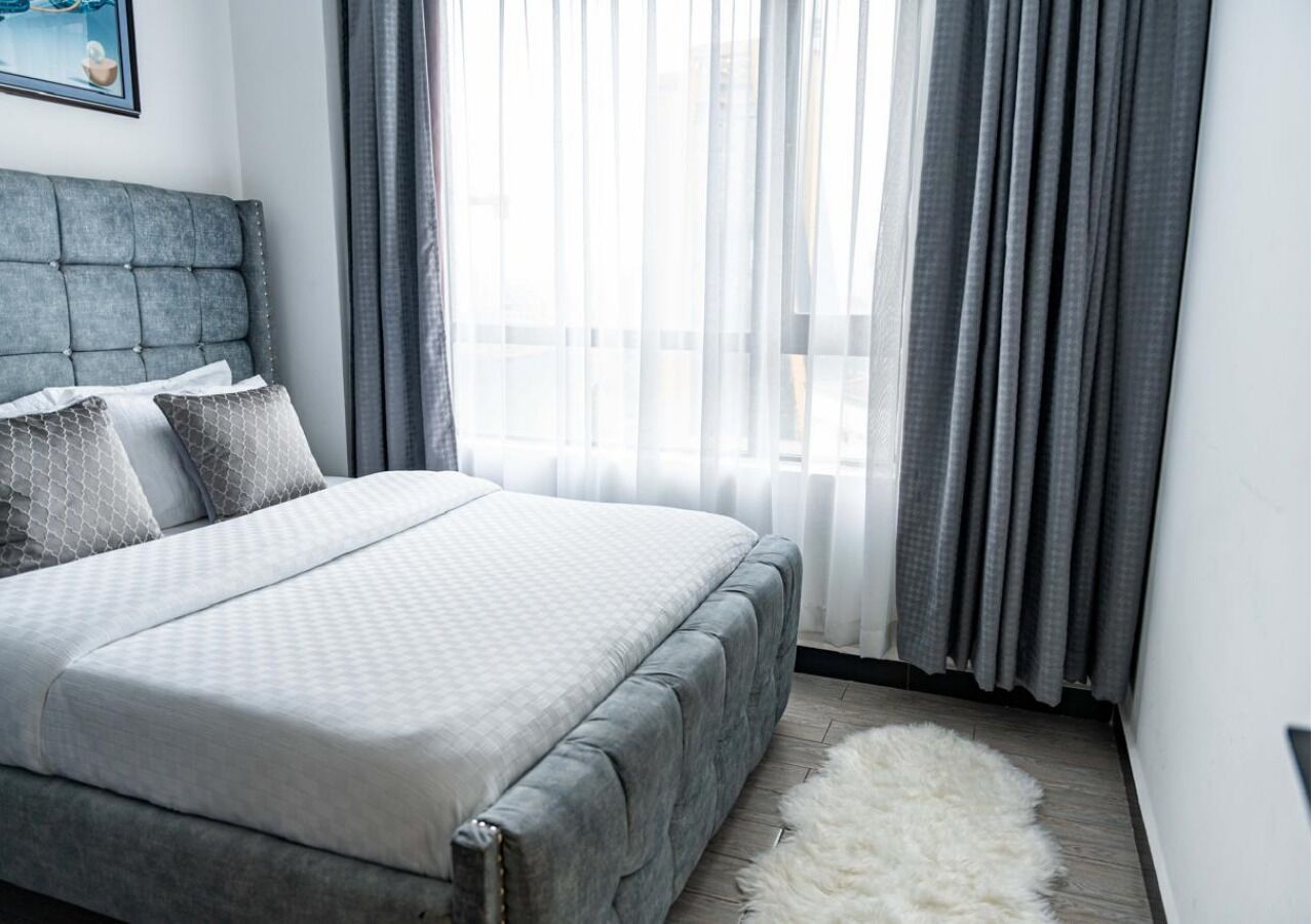 A bedroom with a bed, grey curtains, a white furry rug, and a grey headboard.