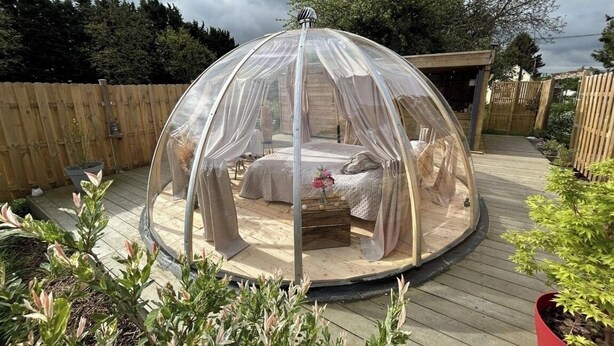 Ein runder, durchsichtiger Pavillon mit einem Bett und einem kleinen Tisch, umgeben von einer Holzterrasse und Pflanzkübeln.