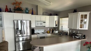 A modern kitchen with white cabinetry, stainless steel appliances, a central island, and a window with blinds. - Cozy Snowbird Retreat on Beautiful Lake June. Monthly Winter Rates (Lake Placid)