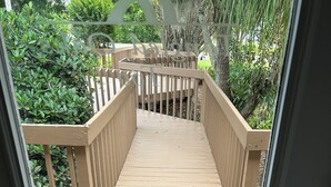 A wooden deck leading to a covered outdoor area with a view of greenery. - Cozy Snowbird Retreat on Beautiful Lake June. Monthly Winter Rates (Lake Placid)