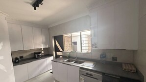 A modern kitchen with white cabinets, a built-in dishwasher, a window with a view of a brick wall, and a ceiling-mounted light fixture. - Serene 3-bedroom House Rental in Secure Kempton Park Complex (Kempton Park)