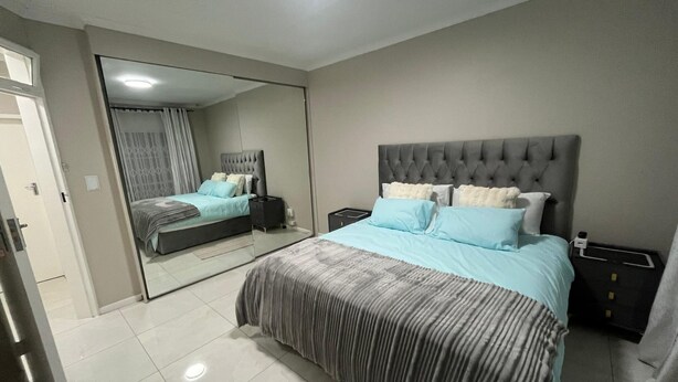 A bedroom with a large bed, a mirror, a nightstand, and a wardrobe. - Serene 3-bedroom House Rental in Secure Kempton Park Complex (Kempton Park)
