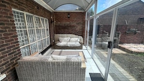 A covered patio with a wicker sofa, a glass door, and a brick wall. - Serene 3-bedroom House Rental in Secure Kempton Park Complex (Kempton Park)