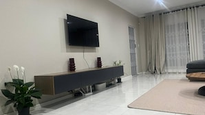 A modern living room with a flat-screen TV mounted on the wall, a dark-colored console table with decorative items, a potted plant, and a large window with sheer curtains. - Serene 3-bedroom House Rental in Secure Kempton Park Complex (Kempton Park)