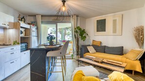 A modern kitchen with a central island, a dining area with a bar stool, a sofa with cushions, and a large window. - Cosy T2 Apartment Facing the Sea with Private Terrace, Tennis Court, and Beach Access (Sète)