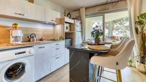 A modern kitchen with a built-in washing machine, a coffee maker, and a dining area with bar stools. - Cosy T2 Apartment Facing the Sea with Private Terrace, Tennis Court, and Beach Access (Sète)