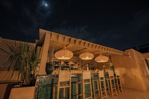 Un área de bar al aire libre con sillas de mimbre, lámparas colgantes, una planta de cactus y una pared de azulejos.