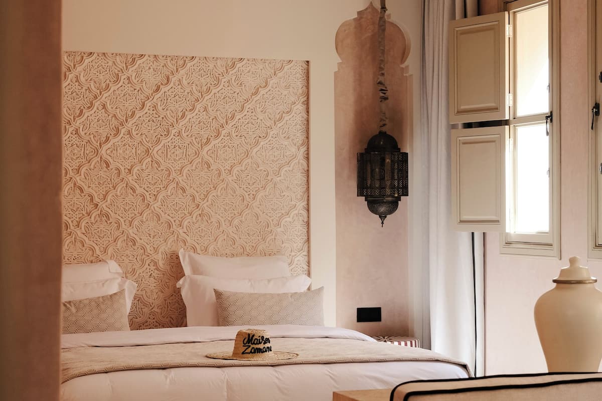 A bedroom with a patterned headboard, a bedside table, a lamp, a mirror, and a window with curtains.