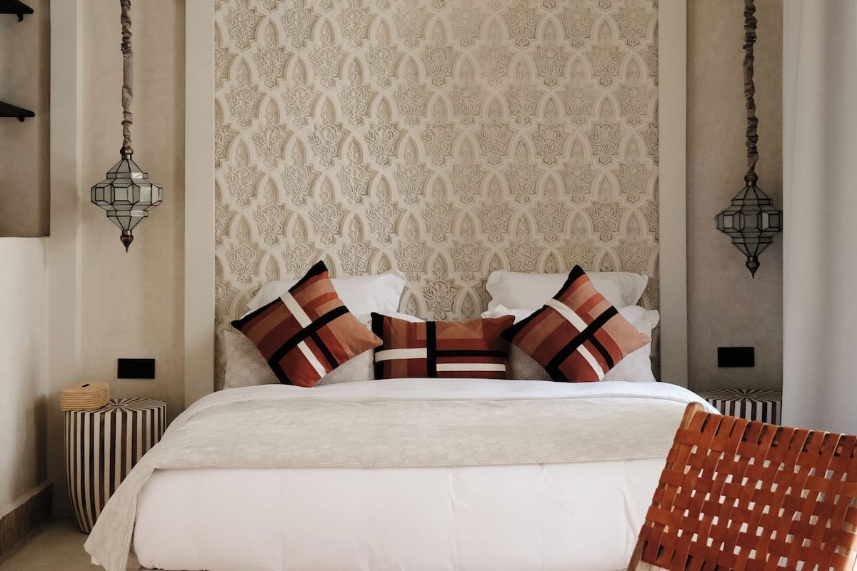 A bedroom with a large bed, two hanging lamps, a wooden chair, and a patterned wall.